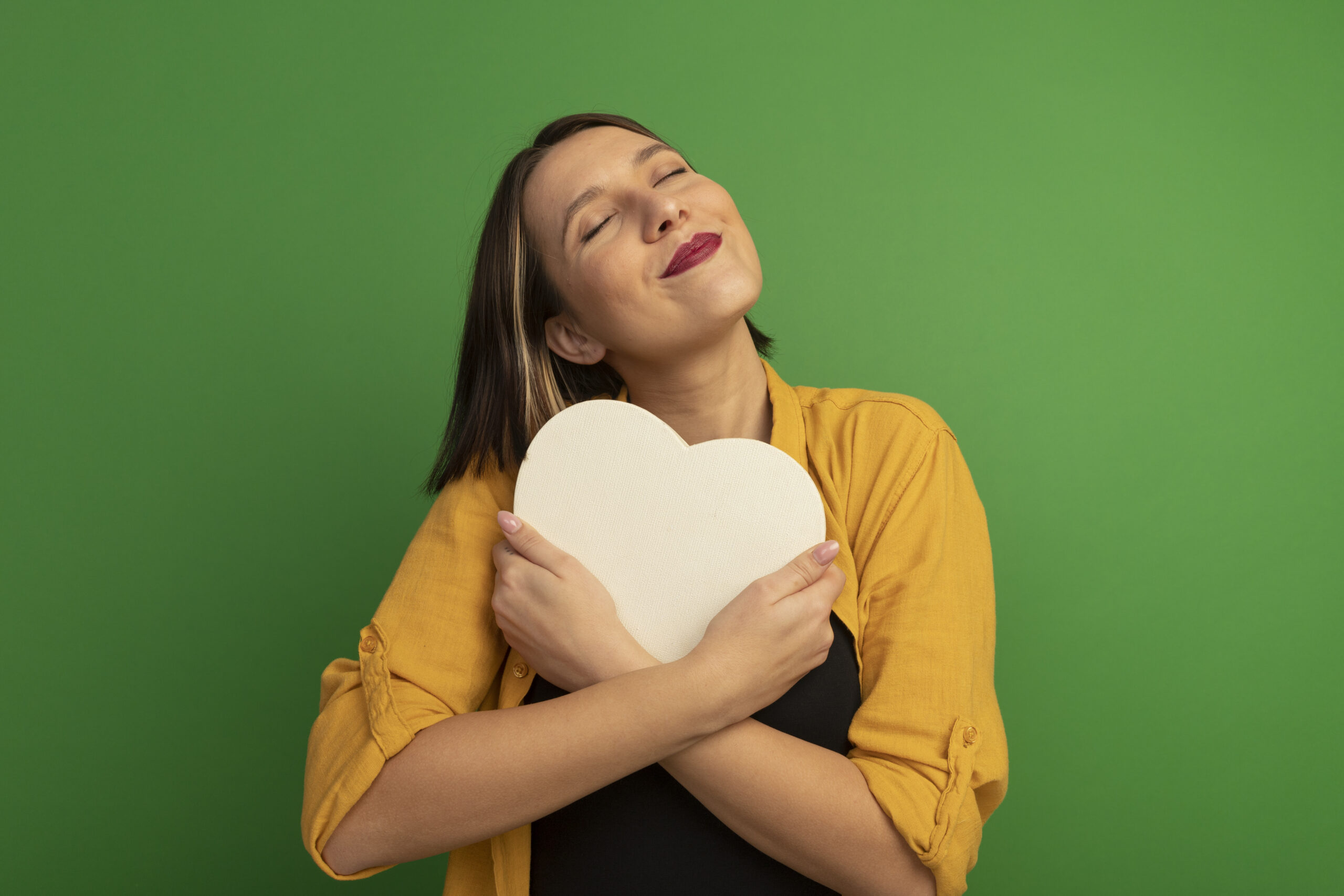pleased pretty caucasian woman hugs heart shape isolated on green background with copy space